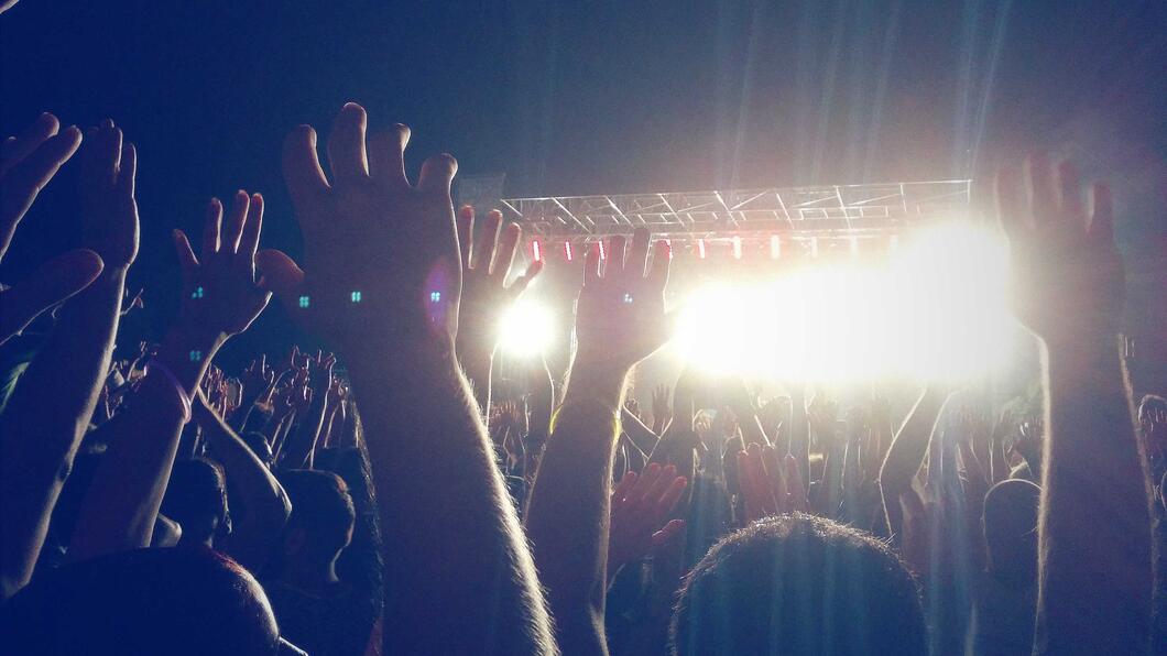 Foto van een buitenfestival in de schemering , in de verte zie je het podium met veel licht en op de voorgrond handen van het publiek die omhoog aan het klappen en juichen zijn