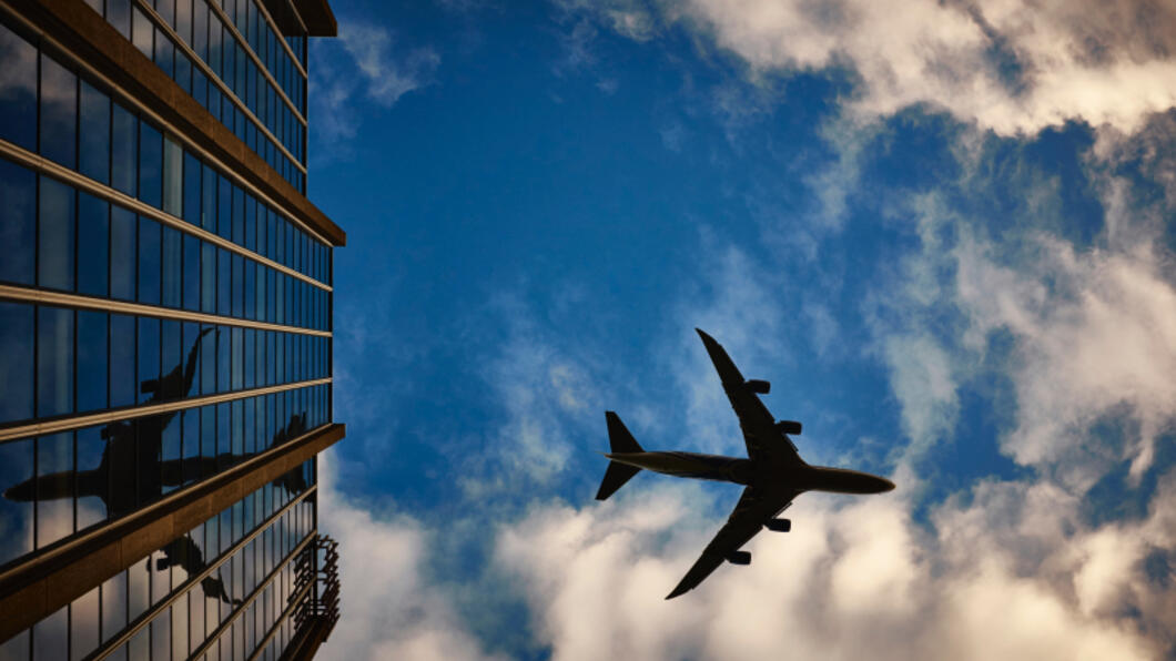 Foto van een vliegtuig tegen een blauwe lucht met wolken en een stuk van een gebouw