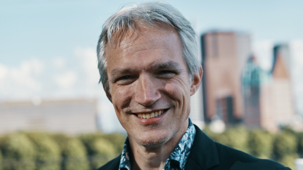 Foto van Berend Potjer die lachend in de camera kijkt. Blauwe lucht, bomen en stukje skyline Den Haag op de achtergrond